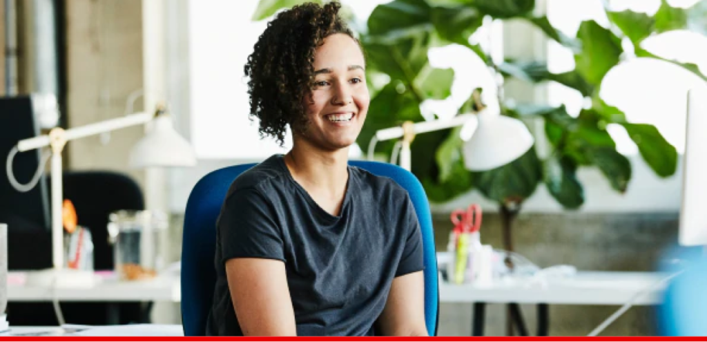 Woman with curly hair in a grey t-shirt sitting in a blue chair, smiling.