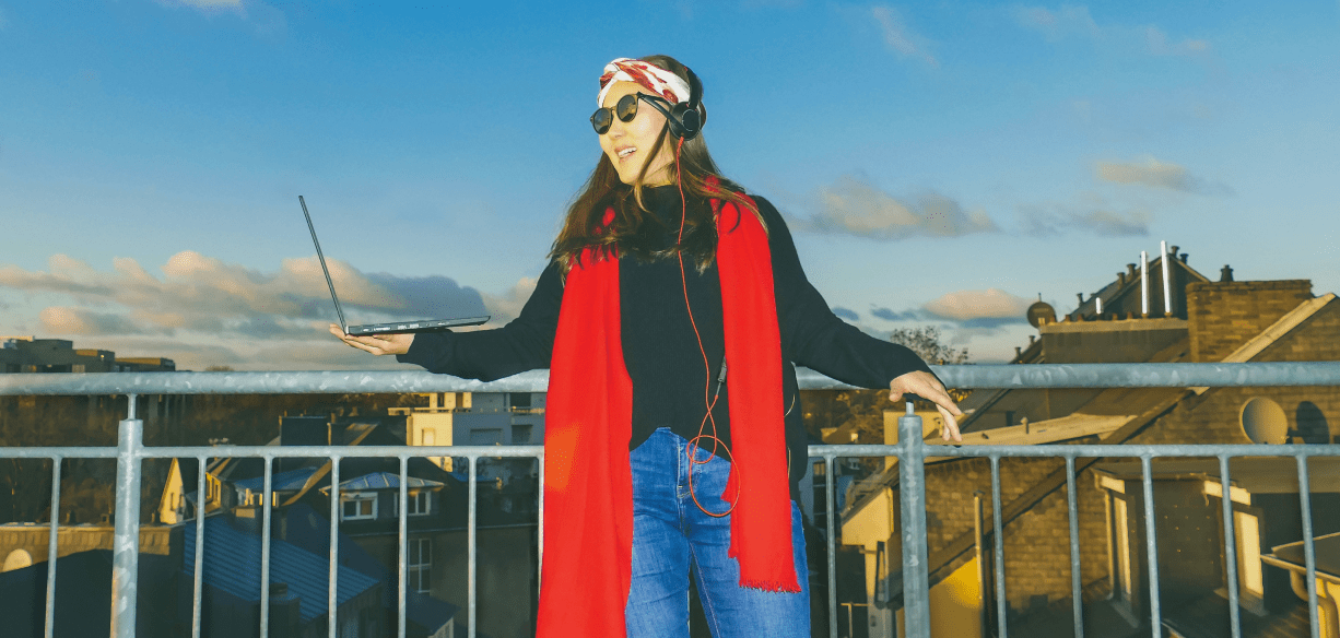 Woman wearing sunglasses and a red scarf, on the balcony with an open laptop.