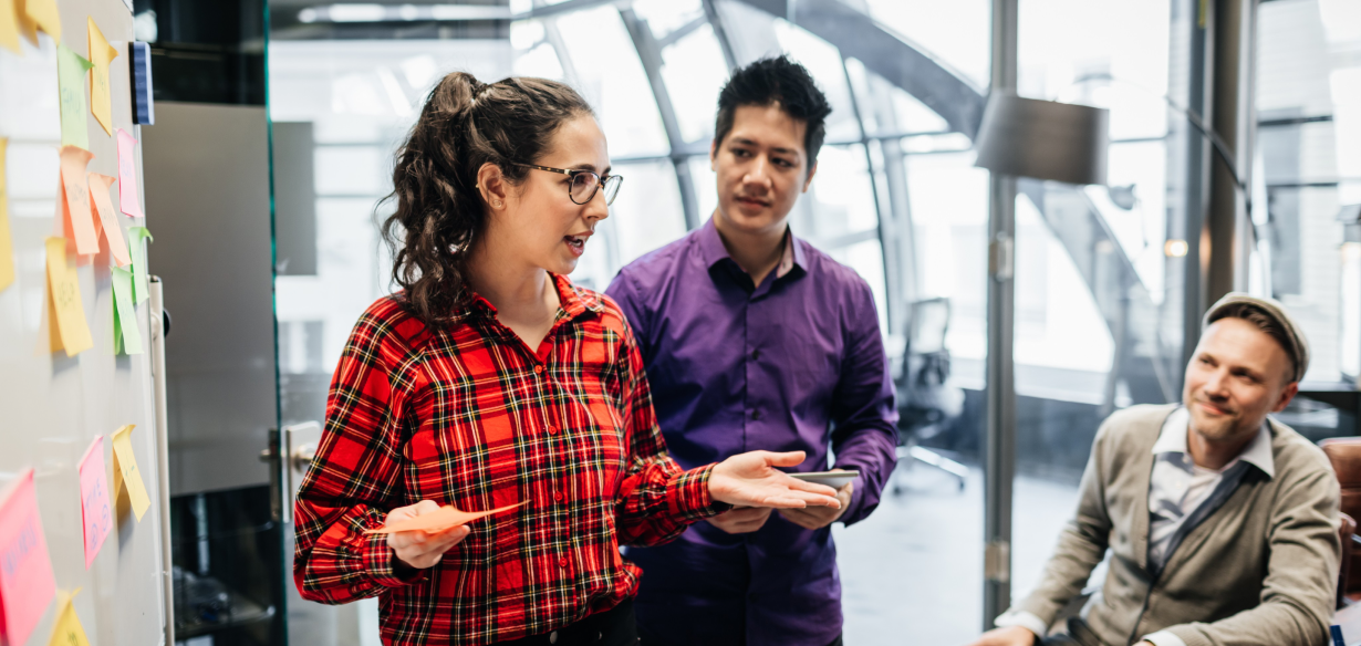 Women in a red check shirt working with two male colleagues in front of a whiteboard with post-it notes.