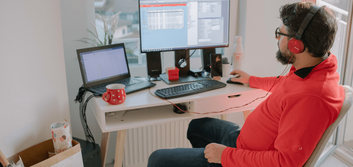 Man in a red jumper wearing headphones, working from home with a large screen and open laptop.