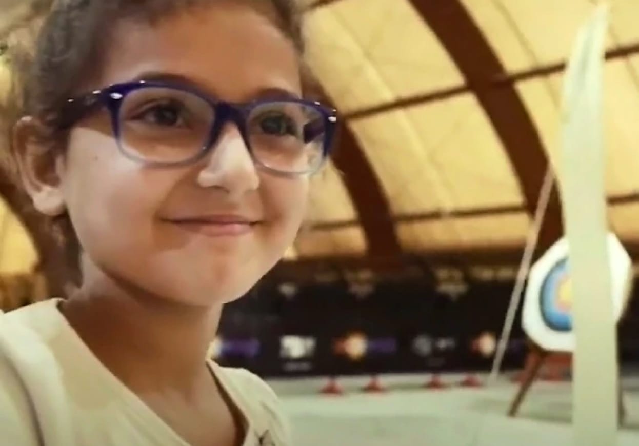 Young girl wearing glasses holding an archery bow standing in front of a target.