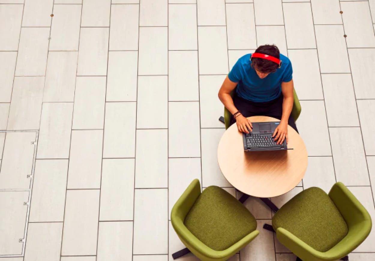 Man sits at a small round table alone opposite two green chairs, with an open laptop.