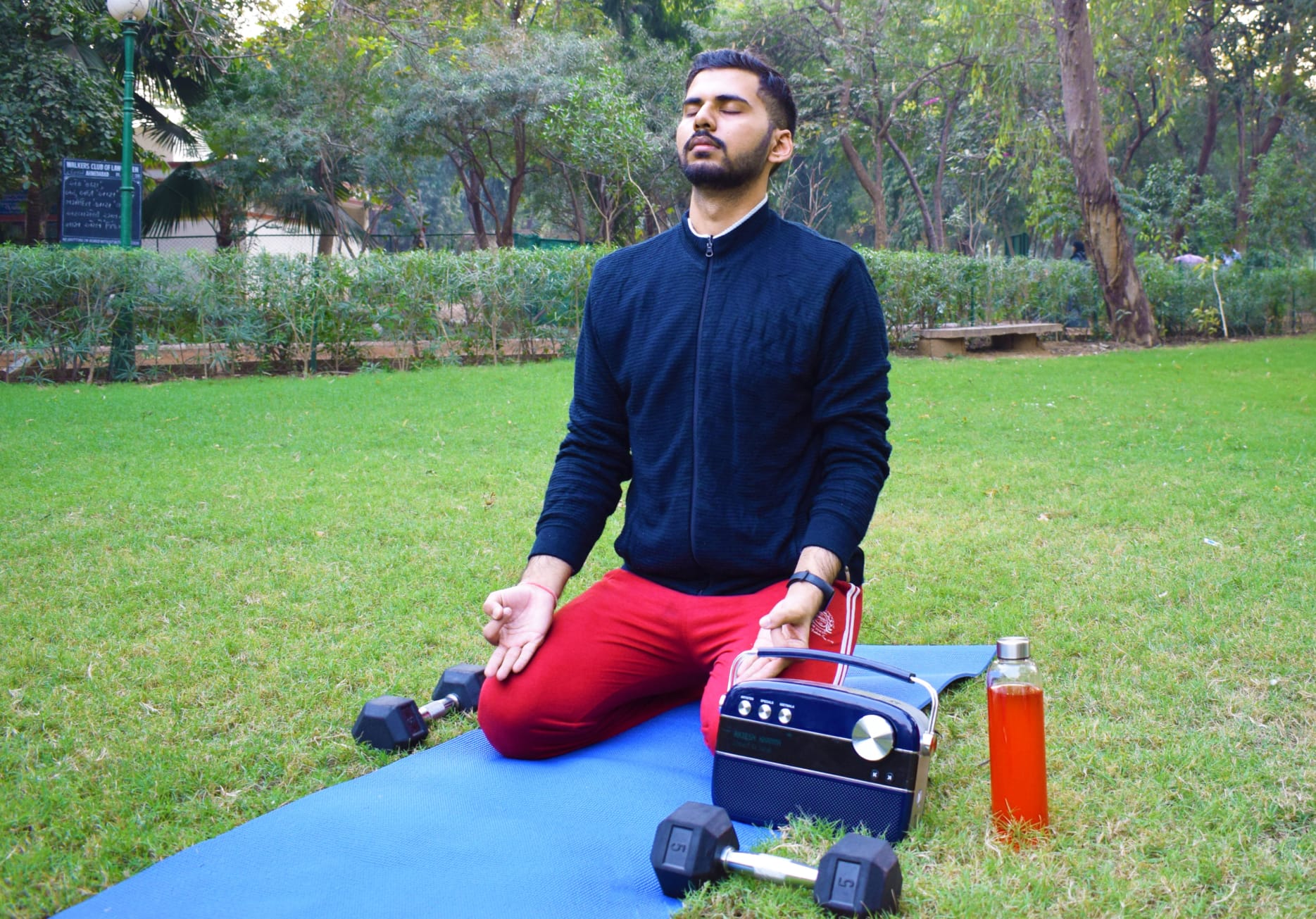 Man kneeling on a blue yoga mat on the grass with his eyes closed next to some weights, a radio speaker and a red water bottle.