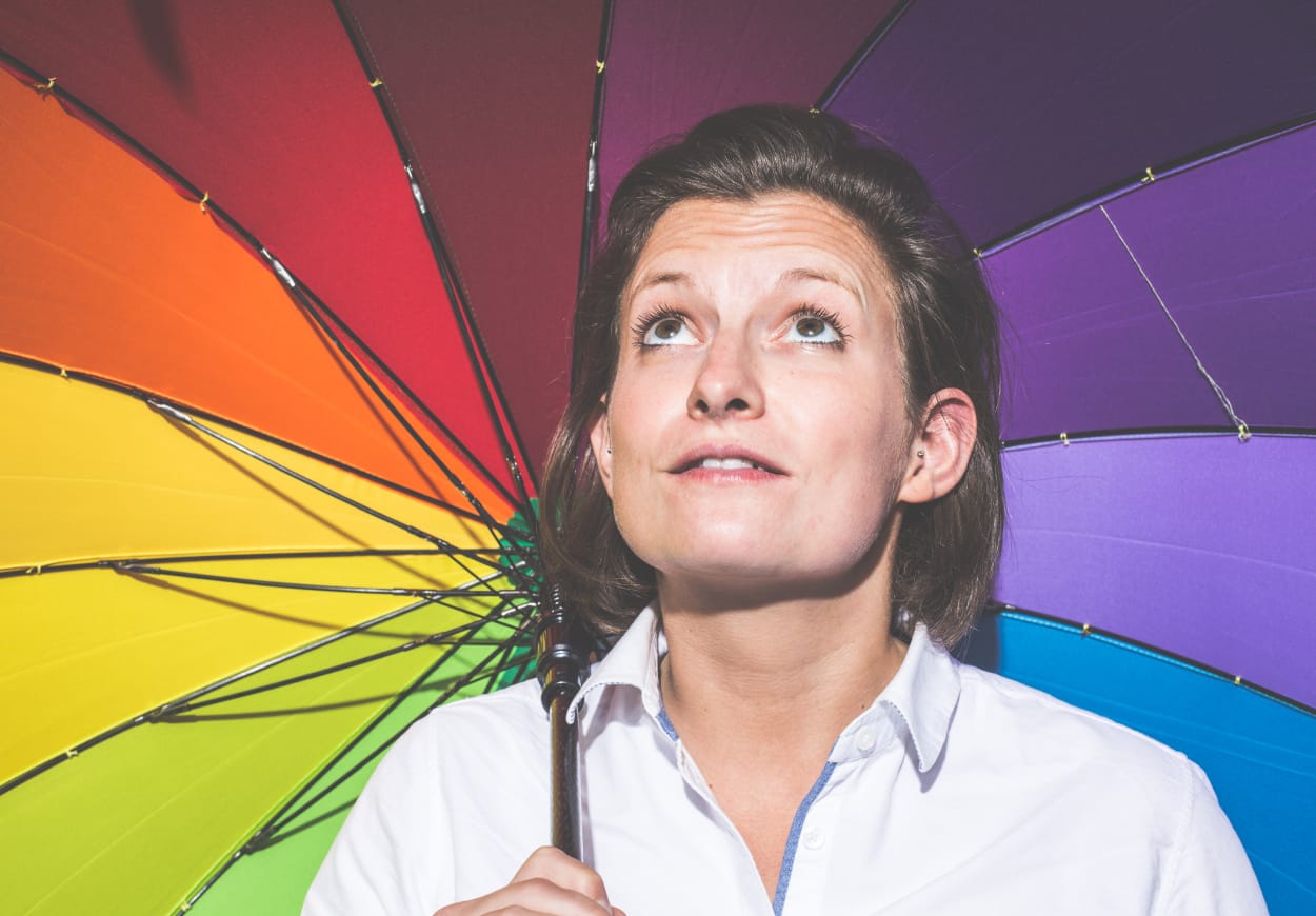 Woman looking up at the sky, whilst holding a rainbow coloured umbrella over her head.