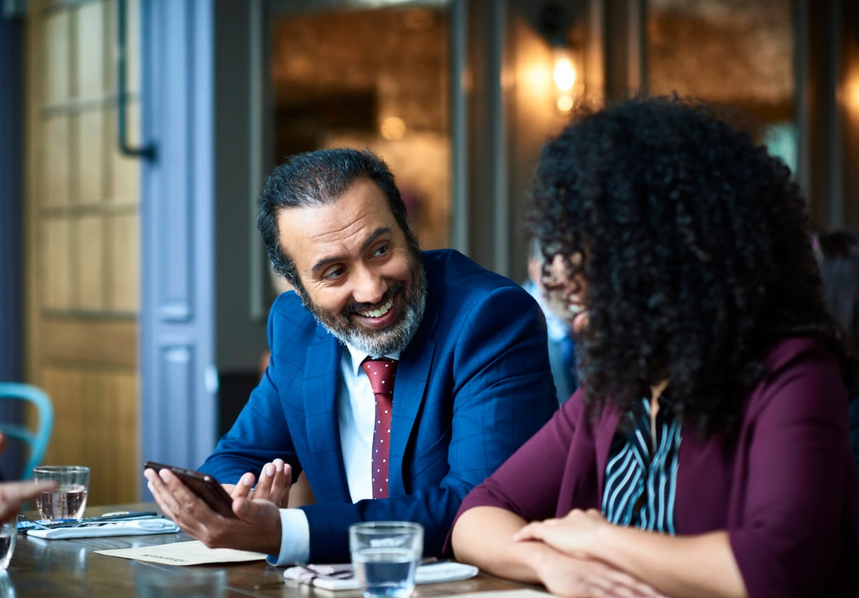 Man with a grey beard and a suit laughs with a woman whilst both are seated at a desk.