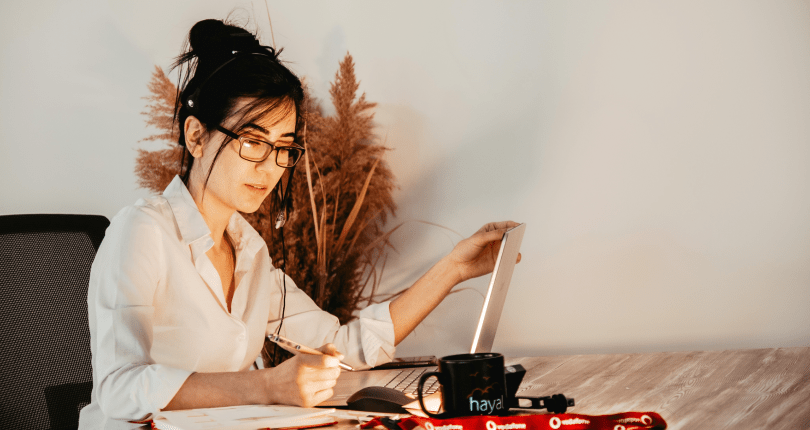 Lady working on laptop with pen and coffee cup.