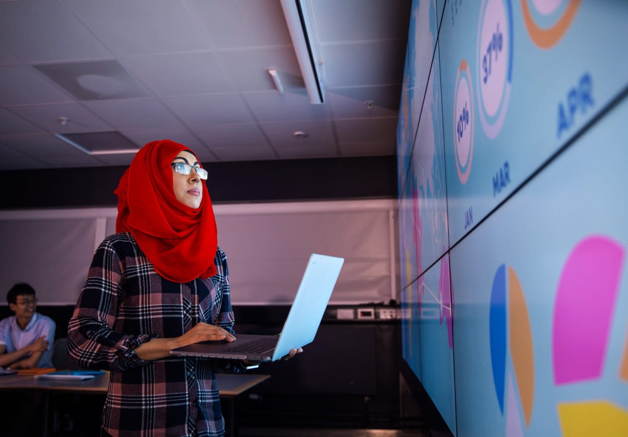 Woman in a red headscarf, working on a laptop in front of a video wall.