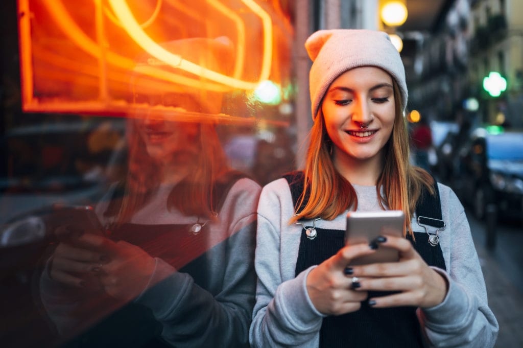 Young woman standing next to neon light using her smartphone at night.