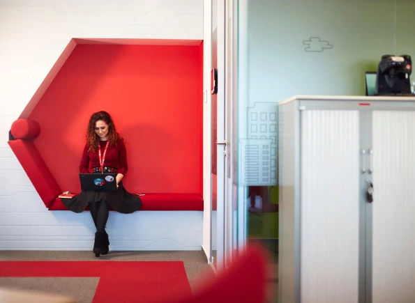 Woman sat on red bench in an office, working on a laptop.